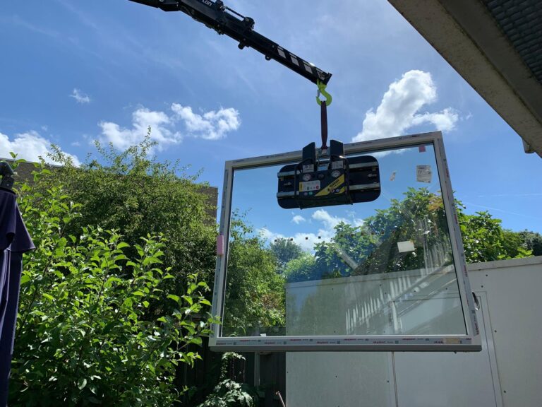 Plaatsing van kunststof kozijn met dubbel glas via vacuüm hijssysteem in tuin onder blauwe lucht.